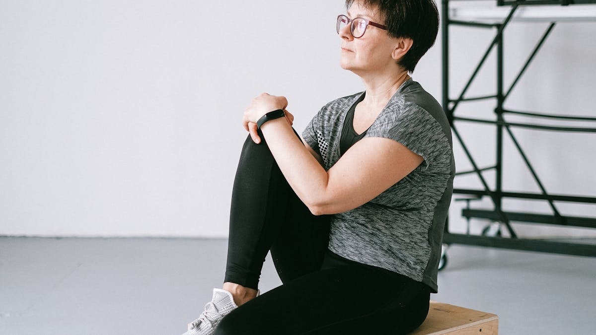 Senior woman in active wear sitting on a wooden box indoors, reflecting on wellness.