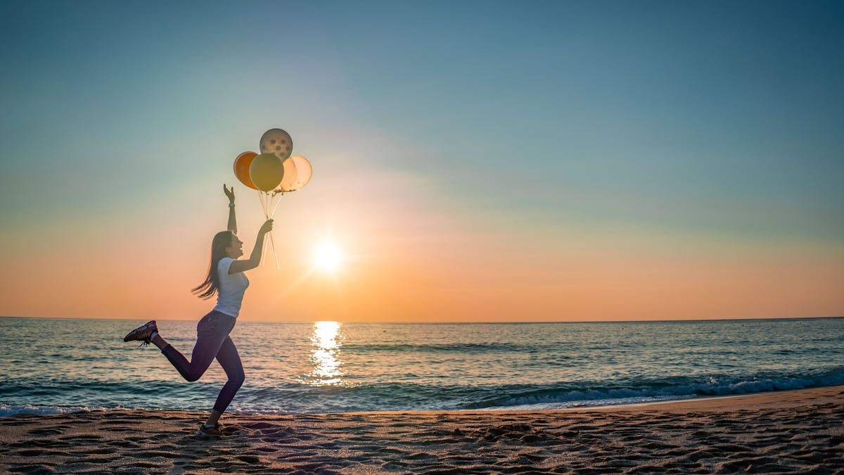 A woman joyfully running with balloons along a beach at sunset, capturing a moment of happiness.